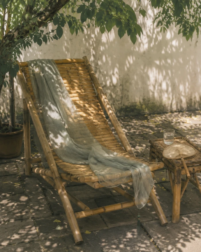 A bamboo lounge chair in summer afternoon shade with a fan and water